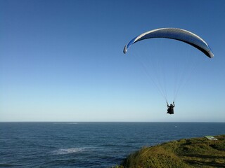 Pessoa voando de paraglider, com mar ao fundo, dia de céu azul, sem nuvens
