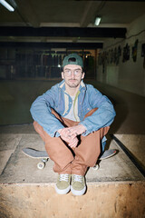 Young Caucasian guy in casual clothes with eyeglasses sitting on skateboard in skatepark, vertical...