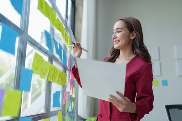 Confident Asian businesswoman Hand holding a pen is thinking while writing on a notepad, notebook to remind in marketing plan information. Online finance, statistics, taxes at the office.