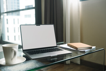 Laptop with white empty screen, notebook and coffee cup on glass table in living room