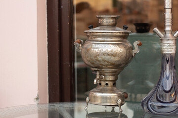 Metal copper bronze old vintage samovar and glass hookah flask stand on table near display window...