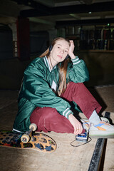 Vertical flash shot of young Caucasian woman in streetwear sitting on floor in skatepark listening...