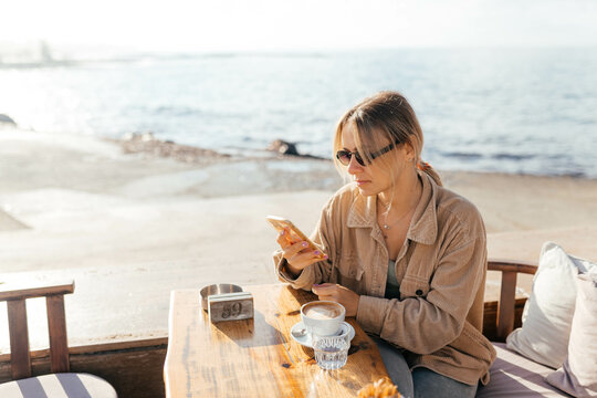 A woman in glasses holding the phone and sitting near the sea in the cafe with a cup of cappuccino ooking relaxed 