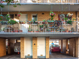 an old brick residential apartment building with cars parked in the carports on the ground floor. Pots of decorative plants on the railing of the balcony.