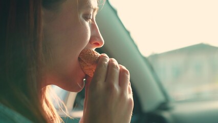 Young woman takes bite of burger while traveling in car. Young woman indulges in quick cuisine....