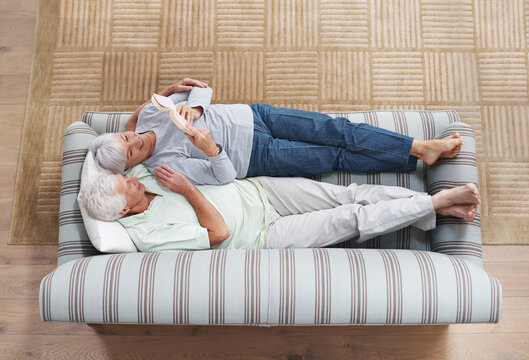 Top view, senior couple and reading book on sofa in living room for story, knowledge and relax in home. Retirement, elderly man and woman lying on couch for love, information and bonding together