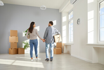 Fototapeta na wymiar Young couple moving into their new home. Back view of happy, loving man an woman holding hands and standing together in spacious empty unfurnished living room in their new house or apartment