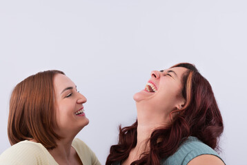 2 young women, close friends, laughing out loud while taking a selfie indoors, with light background. Concept of friendship and fun