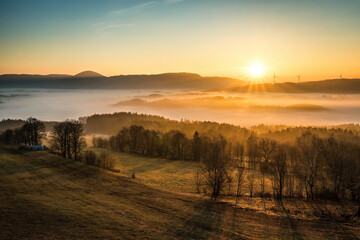 The sun illuminates a valley with thick inversion fog, wind farms and the Královecký špičák...