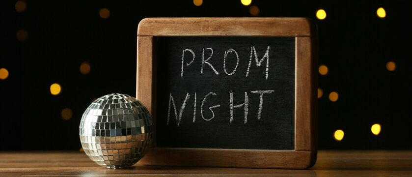 Chalkboard with text PROM NIGHT and disco ball on wooden table against blurred light