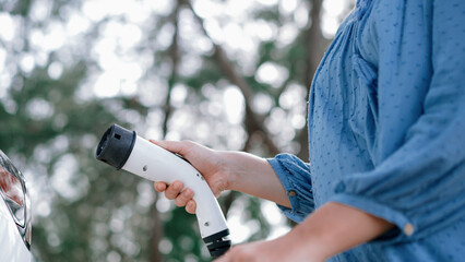 Closeup woman recharge EV electric car's battery at parking lot in natural green park. Clean energy...