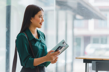 Elegant Asian business professional standing by the window, holding a clipboard in a bright office space.