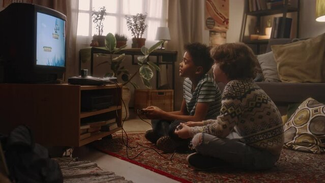 Side full handheld shot of two diverse best friends sitting on carpeted floor and cheering at passing level in old-school arcade video game at retro-styled interior, atmosphere of nostalgic 90s
