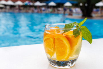 Citrus mint leaves infused water, cocktail, lemonade in glass near pool. Summer iced cold drink with lemon, orange, mint on blue water white marble background, copy space, side view