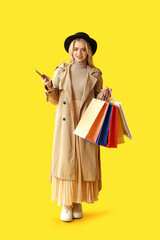 Young woman with modern mobile phone and shopping bags on yellow background