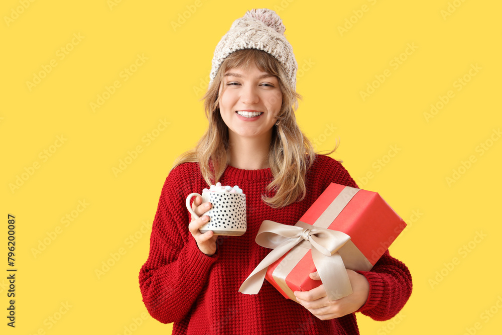 Poster Pretty young woman holding cup of hot chocolate and Christmas gift box on yellow background