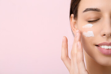 Beautiful young woman applying face cream on pink background