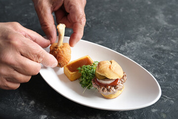 Hands arrange a gourmet fast food course of a mini burger, fried potato square with ketchup and a chicken drumstick on a white plate, copy space, selected focus