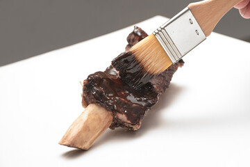 Short rib is coated with flavored glaze using a brush on a white board, preparation for a tasty barbeque, cooking concept, copy space, selected focus