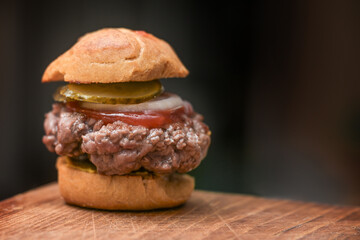 Mini burger with bun, mustard, beef patty, ketchup, onion and gherkin on a rustic wooden kitchen board against a dark background, copy space, selected focus
