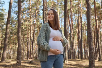 Attractive smiling brown haired Caucasian pregnant woman relaxing in spring forest expectant mother...