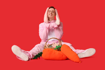 Happy young man in Easter bunny costume sitting near plush carrots and basket on red background