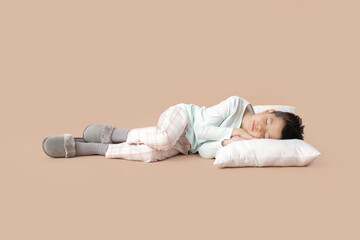 Cute little Asian boy in pajamas with pillow sleeping on beige background