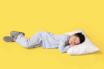 Cute little Asian boy in pajamas with pillow sleeping on yellow background