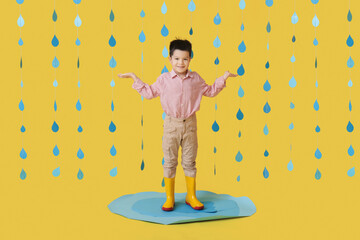 Cute little Asian boy with gumboots and raindrops standing in paper puddle on yellow background