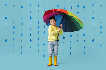 Cute little Asian boy in gumboots with umbrella and paper raindrops on blue background