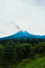 Mount Merapi in Indonesia in the afternoon is very clear and majestic against a background of blue sky and green forest with a ravine flowing cold lava in the foreground.