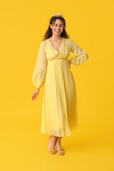 Beautiful young African-American woman in stylish prom dress and tiara on yellow background