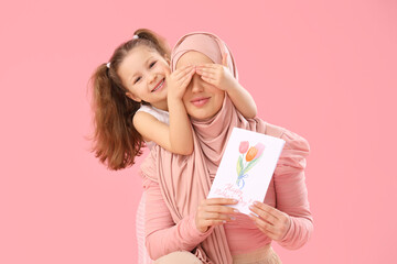 Little girl covering her Muslim mother's eyes with card on pink background