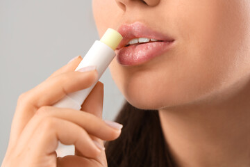 Young woman applying lip balm on grey background, closeup
