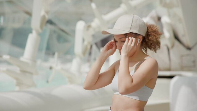 An active young woman in a white sports bra adjusts her cap, getting ready to continue her workout in a modern urban area with distinct architectural design. Slow motion. 
