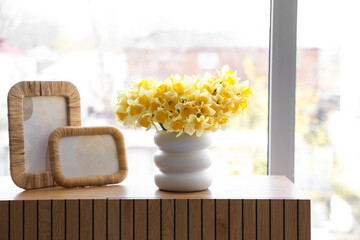 Vase with daffodils and frames on commode near window in room