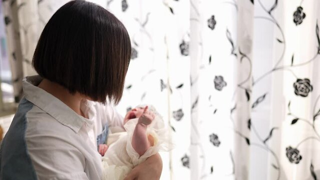 ２０代の台湾人の母親が生後１ヶ月の女の子の赤ちゃんを室内のカーテンの前であやしている風景 A Taiwanese mother in her 20s is torting a baby girl who is 1 month old in front of an indoor curtain.