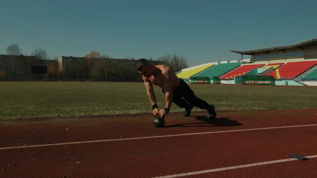 Young man is training at city stadium and doing push-ups on kettlebell and overhead swings. Effective exercise for training explosive strength of muscles of hamstrings and buttocks