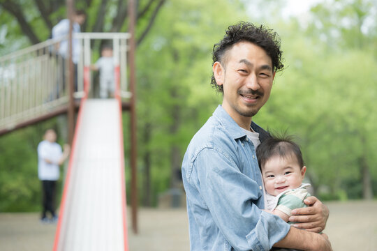 新緑の屋外で赤ちゃんを抱くパパ　イクメンや男性の育児や子育てのイメージ　カメラ目線