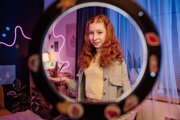 Portrait of stylish teen girl standing in her room posing for camera, through circle lamp medium...