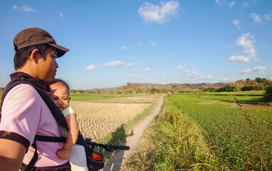 Asian man is holding a baby while cycling and taking selfies with a backdrop of hills and rice fields when the weather is sunny in the afternoon.