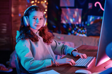Portrait of confident Caucasian girl wearing hoodie and headphones sitting at desk in her room...