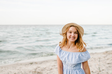 Fototapeta na wymiar Happy beautiful girl standing on beach ocean and enjoying sunny summer day on vacation. Young smiling blond woman in straw hat walks near sea and looks at camera. Portrait of stylish female on sand.
