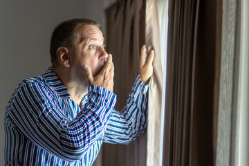 Shocked man in striped pajamas looks out behind curtained window