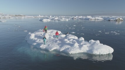 In the tranquil, icy waters of Antarctica, a team of explorers gathers on a floating ice floe, surrounded by snow-covered islands, under the bright expanse of a cloudless sky.