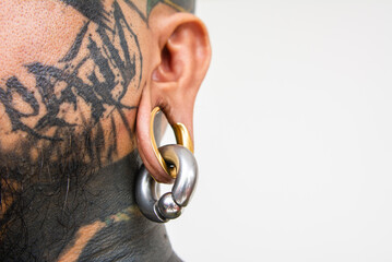 Closeup shot of a gold flesh tunnel, also known as a earplug, earspool. Large gauge plug on the earlobe of a man with face tattoos. - obrazy, fototapety, plakaty