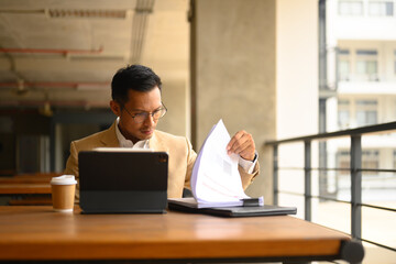 Serious busy businessman reading financial documents and working with digital tablet