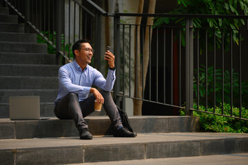 Cheerful male office worker sitting on stairs with his laptop in the city and using mobile phone - 796365426