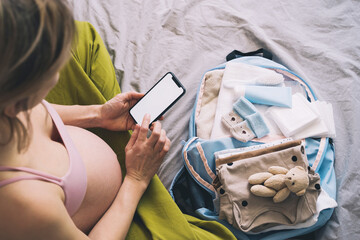 Pregnant woman getting ready for labor packing stuff for hospital, making notes or checklist in smartphone. Baby clothes, necessities for mother and newborn in maternity bag.
