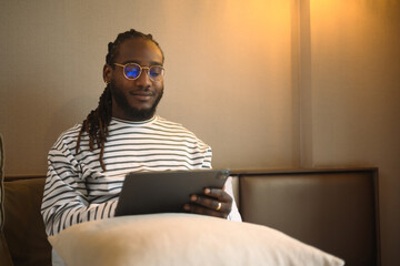 Happy young African American man sitting on bed using digital tablet